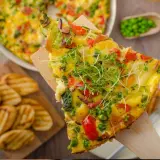 Rebanada de frittata de verduras con guisantes, pimientos y brotes frescos, servida con patatas a la parrilla.