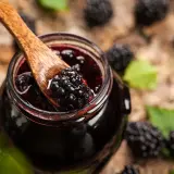 Cuchara de madera recoge mermelada de moras silvestres de un tarro, rodeado de moras frescas y hojas verdes.
