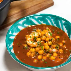 Un tazón de garbanzos con langostinos en una salsa rojiza, adornado con perejil picado. Un plato reconfortante y lleno de sabor.