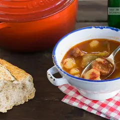 Un plato humeante de patatas a la riojana, con chorizo y pimientos, acompañado de pan rústico y una olla roja.