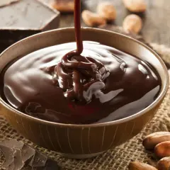 Un chorro de ganache de chocolate cae en un cuenco, rodeado de trozos de chocolate y granos de cacao.