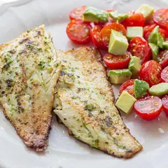 Dos filetes de lubina a la plancha con hierbas, acompañados de una ensalada fresca de tomates cherry y aguacate en cubos.