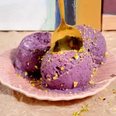 Tres bolas de helado casero de arándanos con pistachos picados, servidas en un plato rosa. Una cuchara dorada se hunde en el helado.