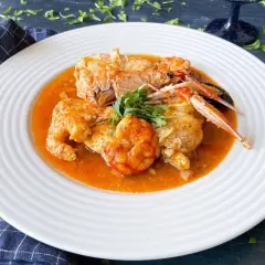 Un plato de rap a la marinera con gambas y cigalas en una salsa rojiza, decorado con perejil fresco.
