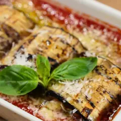 Delicioso pastel de berenjenas gratinado con queso y bañado en salsa de tomate, adornado con hojas de albahaca fresca.