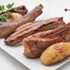 Costillas de cerdo jugosas y doradas, servidas en una bandeja blanca con patatas asadas y una ramita de perejil.