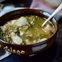 Un cuenco de barro con la palabra "Gallego" escrita, lleno de un reconfortante caldo gallego con patatas, grelos y trozos de pan.
