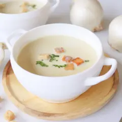 Crema de cebolla cremosa, adornada con picatostes dorados y perejil fresco. Cebollas blancas enteras y cortadas a medias rodean los cuencos.