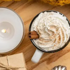 Taza de bebida caliente con abundante **montar nata**, canela y una rama de canela. Una vela encendida y un regalo completan la escena acogedora.