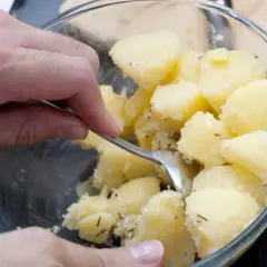 Manos mezclando patatas cocidas con hierbas en un bol de cristal.