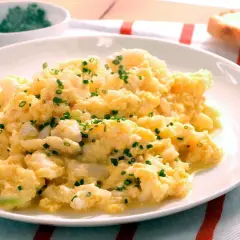 Revuelto de bacalao cremoso, espolvoreado con cebollino fresco, servido en un plato blanco con una rebanada de pan tostado al lado.