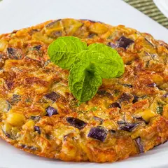 Tortilla de berenjena dorada y esponjosa, con trozos morados de berenjena y patata, decorada con hojas verdes de menta.