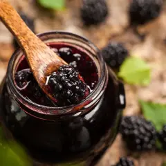Cuchara de madera recoge mermelada de moras silvestres de un tarro, rodeado de moras frescas y hojas verdes.