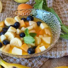 Deliciosa macedonia de frutas con plátano, arándanos, melocotón y manzana en un cuenco de cristal.
