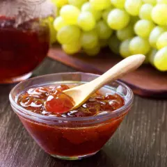 Un cuenco de cristal con mermelada de uvas casera y una cuchara de madera, junto a un racimo de uvas verdes.