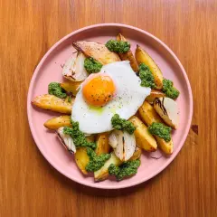 Patatas al horno doradas con huevo frito, cebolla asada y pesto verde, servidas en un plato rosa sobre una mesa de madera.