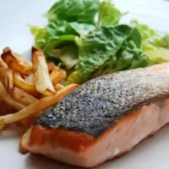 Un jugoso salmón a la plancha con piel crujiente, acompañado de patatas fritas doradas y una fresca ensalada verde.