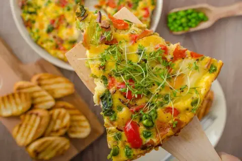 Rebanada de frittata de verduras con guisantes, pimientos y brotes frescos, servida con patatas a la parrilla.