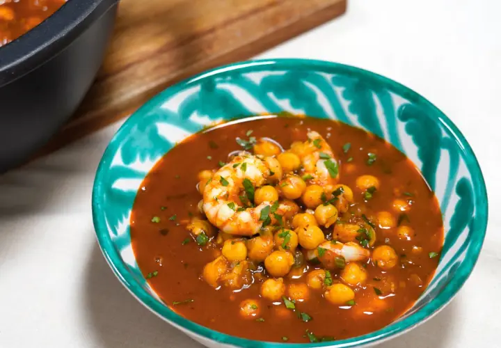 Un tazón de garbanzos con langostinos en una salsa rojiza, adornado con perejil picado. Un plato reconfortante y lleno de sabor.