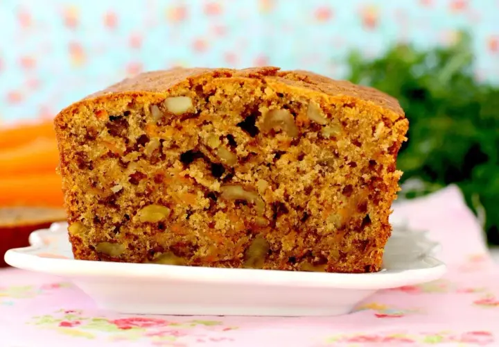 Un delicioso bizcocho de zanahoria, con trozos de nueces y un color dorado apetitoso, servido en un plato blanco sobre un mantel floral.