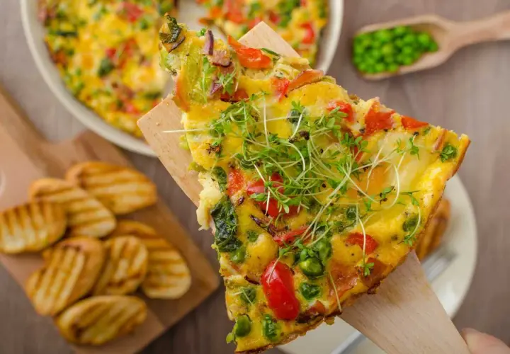 Rebanada de frittata de verduras con guisantes, pimientos y brotes frescos, servida con patatas a la parrilla.