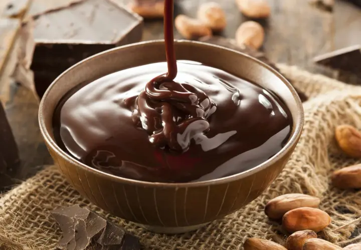 Un chorro de ganache de chocolate cae en un cuenco, rodeado de trozos de chocolate y granos de cacao.