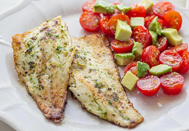 Dos filetes de lubina a la plancha con hierbas, acompañados de una ensalada fresca de tomates cherry y aguacate en cubos.