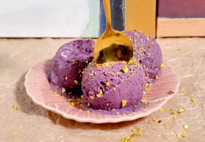 Tres bolas de helado casero de arándanos con pistachos picados, servidas en un plato rosa. Una cuchara dorada se hunde en el helado.