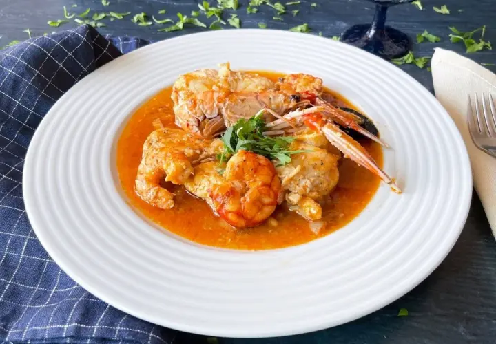 Un plato de rap a la marinera con gambas y cigalas en una salsa rojiza, decorado con perejil fresco.