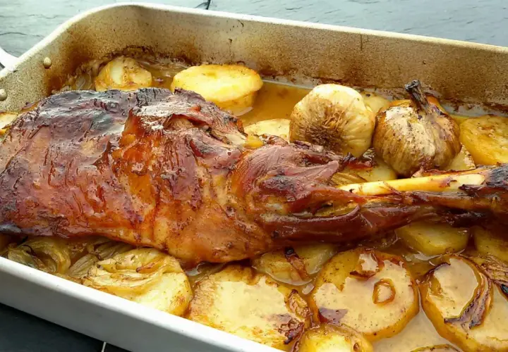 Paletilla de cordero asada con patatas y ajos dorados en una bandeja. Jugosa y apetitosa, lista para servir.