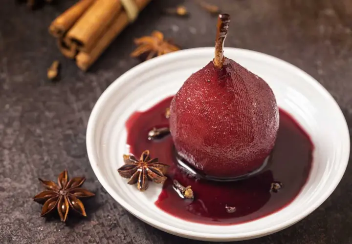 Peras al vino, cocinadas a la perfección, servidas en un plato blanco con especias como anís estrellado y canela.