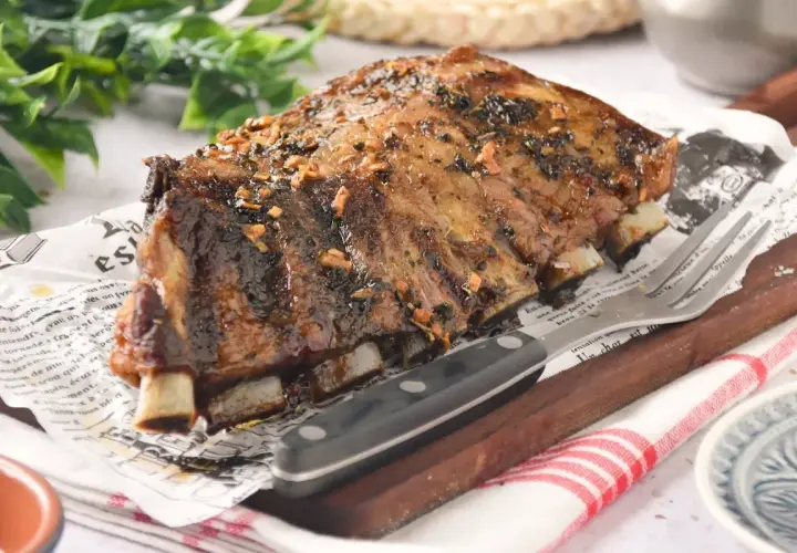 Costillas de churrasco de cerdo glaseadas, servidas sobre papel con texto y acompañadas de un tenedor y cuchillo.