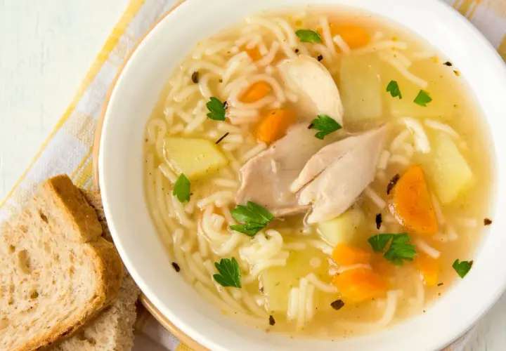 Un reconfortante plato de sopa de pollo receta, con fideos finos, trozos de pollo, patatas y zanahorias, adornado con perejil fresco.