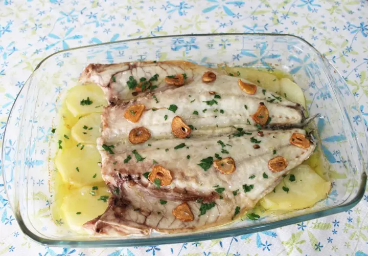 Lubina a la espalda horneada con patatas, ajo y perejil en una fuente de cristal.