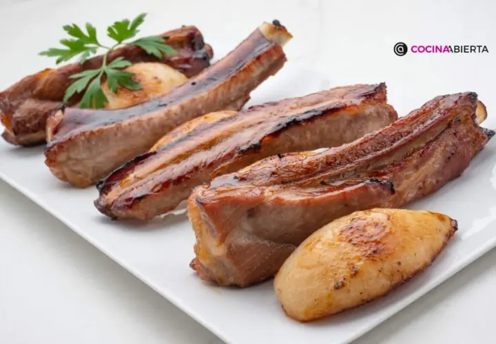 Costillas de cerdo jugosas y doradas, servidas en una bandeja blanca con patatas asadas y una ramita de perejil.