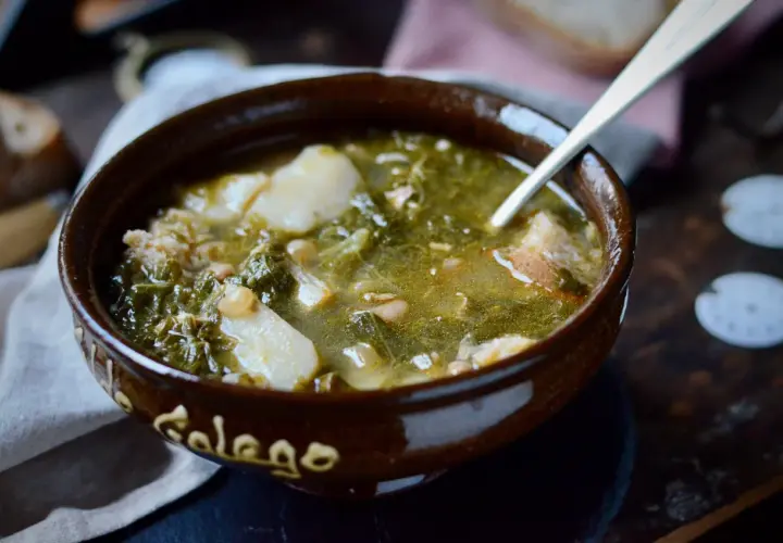 Un cuenco de barro con la palabra "Gallego" escrita, lleno de un reconfortante caldo gallego con patatas, grelos y trozos de pan.