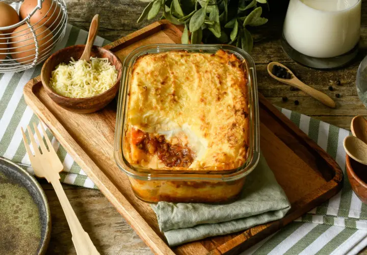 Pastel de carne picada recién horneado, con una capa dorada de puré de patatas y queso gratinado. Se sirve en una fuente de cristal sobre una tabla de madera.