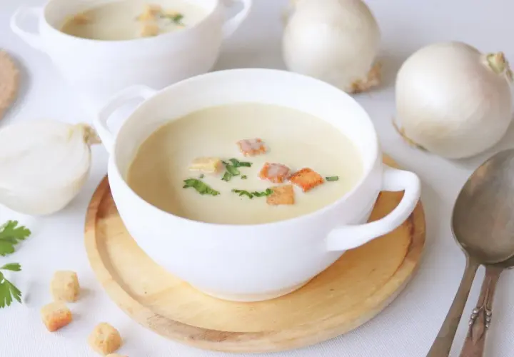 Crema de cebolla cremosa, adornada con picatostes dorados y perejil fresco. Cebollas blancas enteras y cortadas a medias rodean los cuencos.
