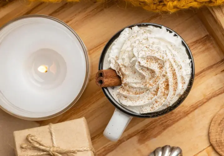 Taza de bebida caliente con abundante **montar nata**, canela y una rama de canela. Una vela encendida y un regalo completan la escena acogedora.