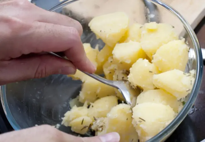 Manos mezclando patatas cocidas con hierbas en un bol de cristal.