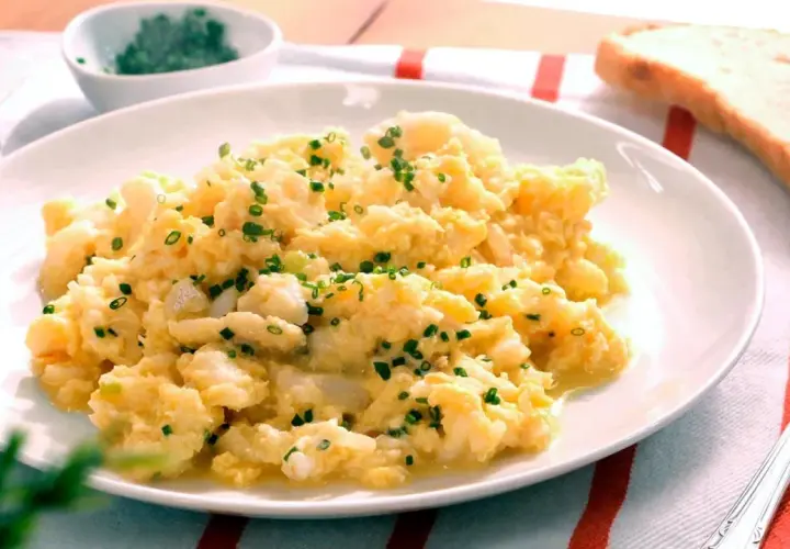 Revuelto de bacalao cremoso, espolvoreado con cebollino fresco, servido en un plato blanco con una rebanada de pan tostado al lado.