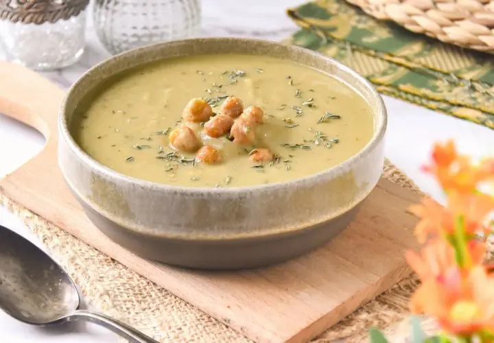 Un tazón de cremosa crema de garbanzos, decorada con garbanzos enteros y hierbas, servida en una tabla de madera.