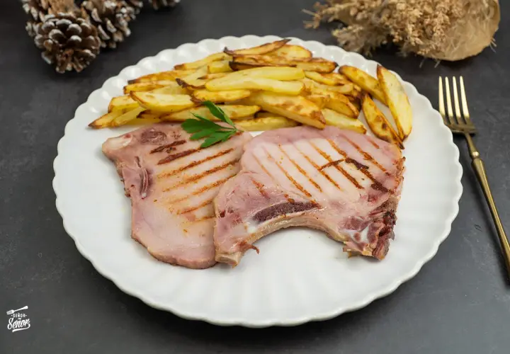 Dos chuletas de sajonia a la parrilla con patatas fritas y perejil en un plato blanco.