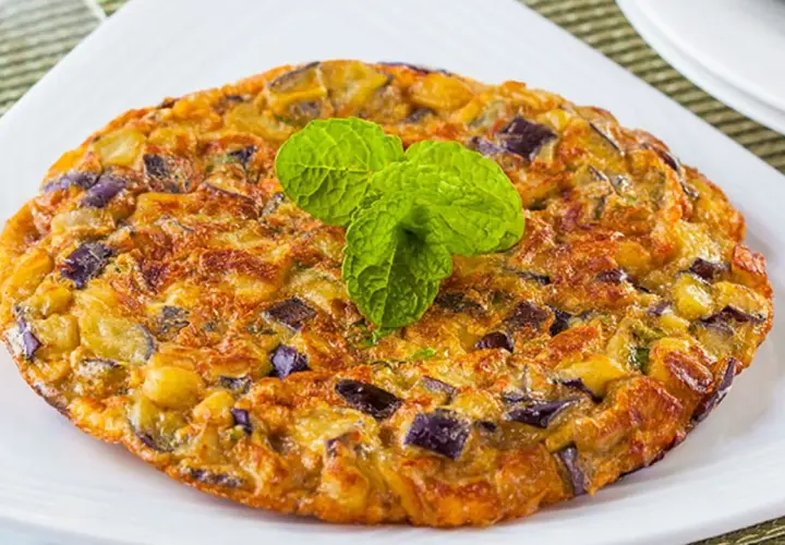 Tortilla de berenjena dorada y esponjosa, con trozos morados de berenjena y patata, decorada con hojas verdes de menta.