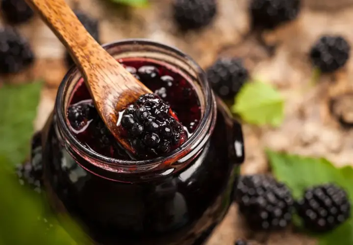 Cuchara de madera recoge mermelada de moras silvestres de un tarro, rodeado de moras frescas y hojas verdes.