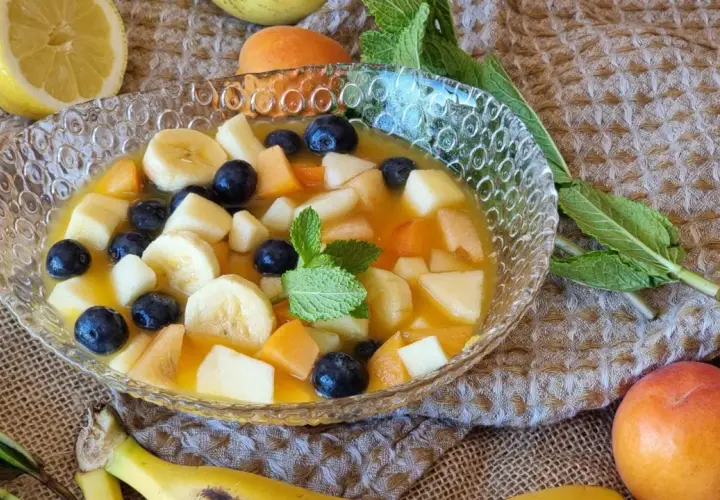 Deliciosa macedonia de frutas con plátano, arándanos, melocotón y manzana en un cuenco de cristal.