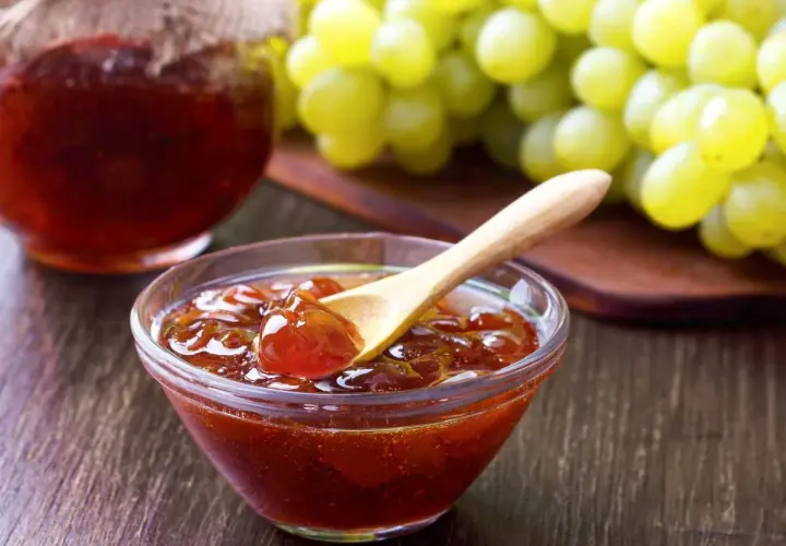 Un cuenco de cristal con mermelada de uvas casera y una cuchara de madera, junto a un racimo de uvas verdes.
