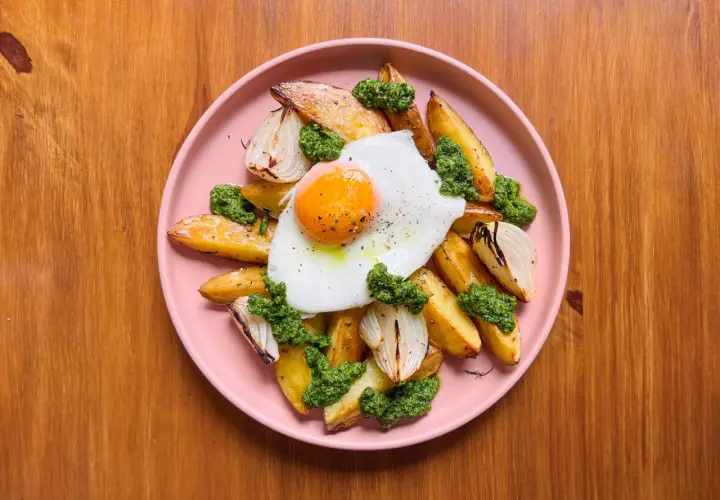 Patatas al horno doradas con huevo frito, cebolla asada y pesto verde, servidas en un plato rosa sobre una mesa de madera.