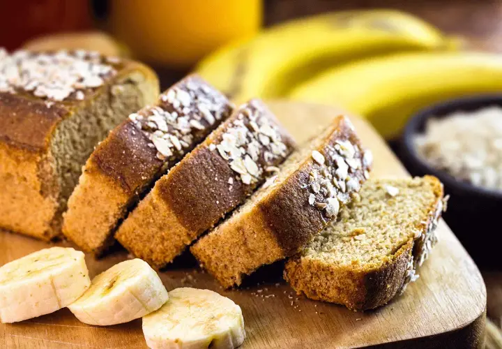 Rebanadas de bizcocho de plátano sin azúcar, cubiertas con avena, junto a plátanos frescos y un tazón de avena.