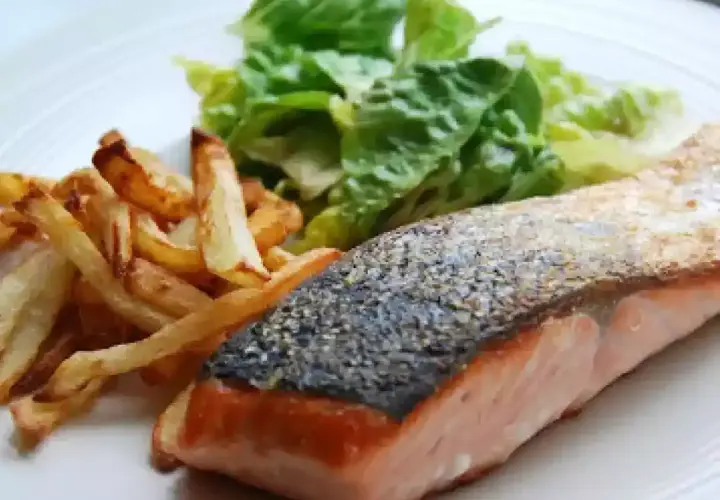 Un jugoso salmón a la plancha con piel crujiente, acompañado de patatas fritas doradas y una fresca ensalada verde.